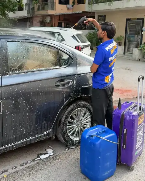 Washer pressure-washing a dark sedan with the water can and purple kit case beside him